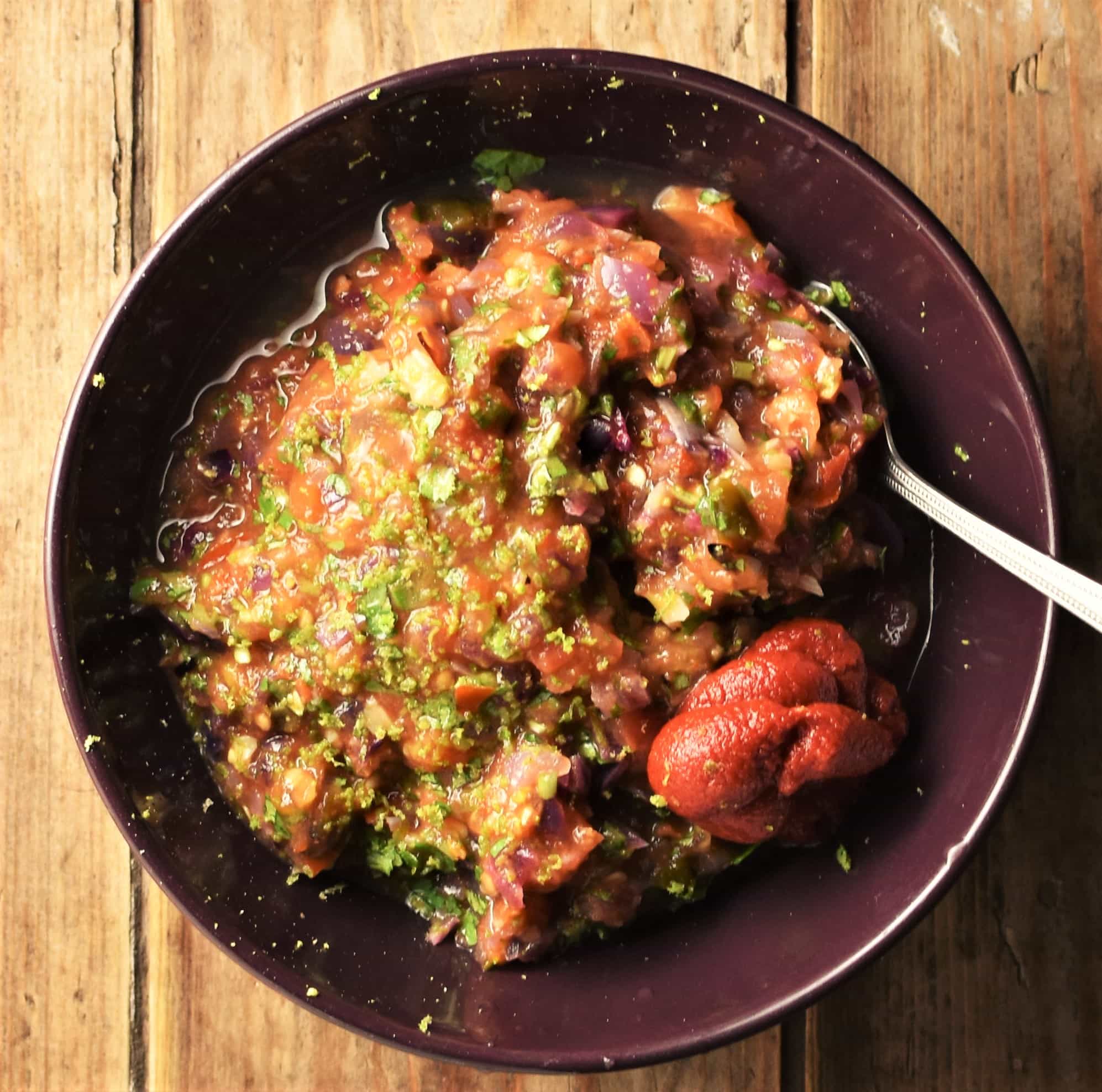 Tomato salsa mixture with tomato paste and lime zest in purple bowl with spoon.