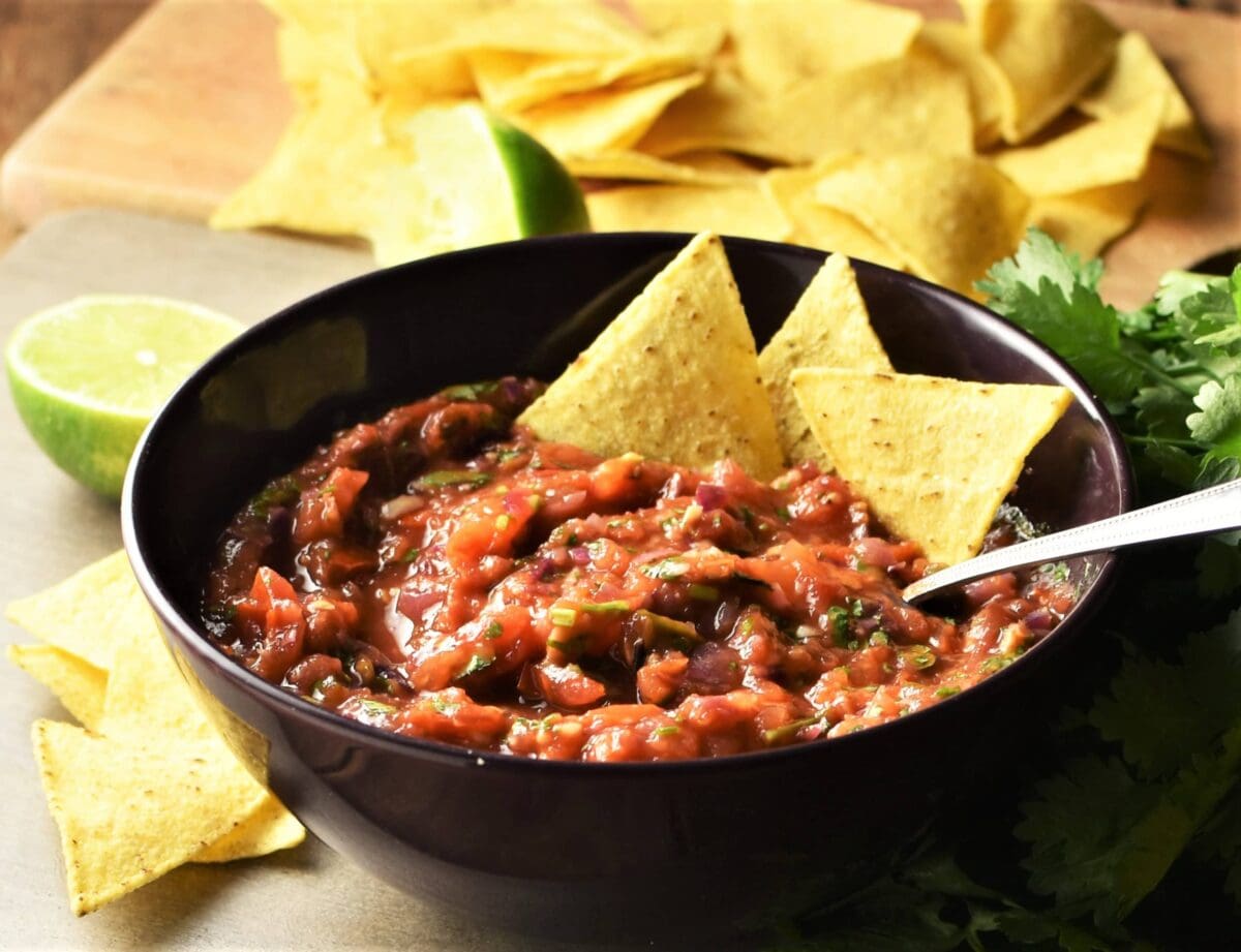 Side view of salsa in purple bowl with nachos and spoon, lime, herbs and nachos in background.