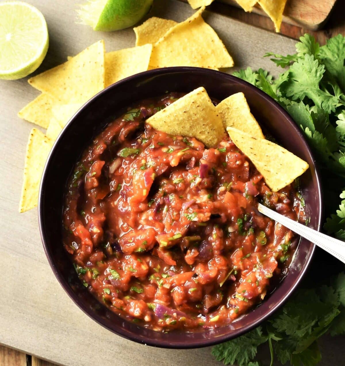 Roasted tomato salsa in bowl with nachos and spoon, nachos, limes and herbs in background.