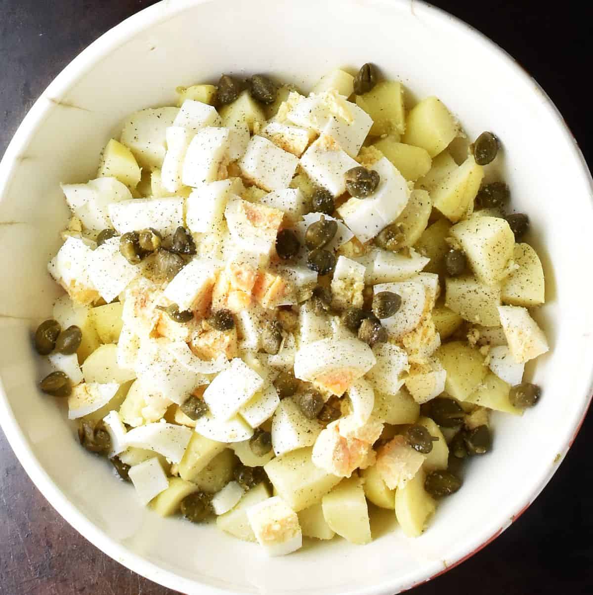 Top down view of cubed potatoes, chopped eggs and capers in white bowl.