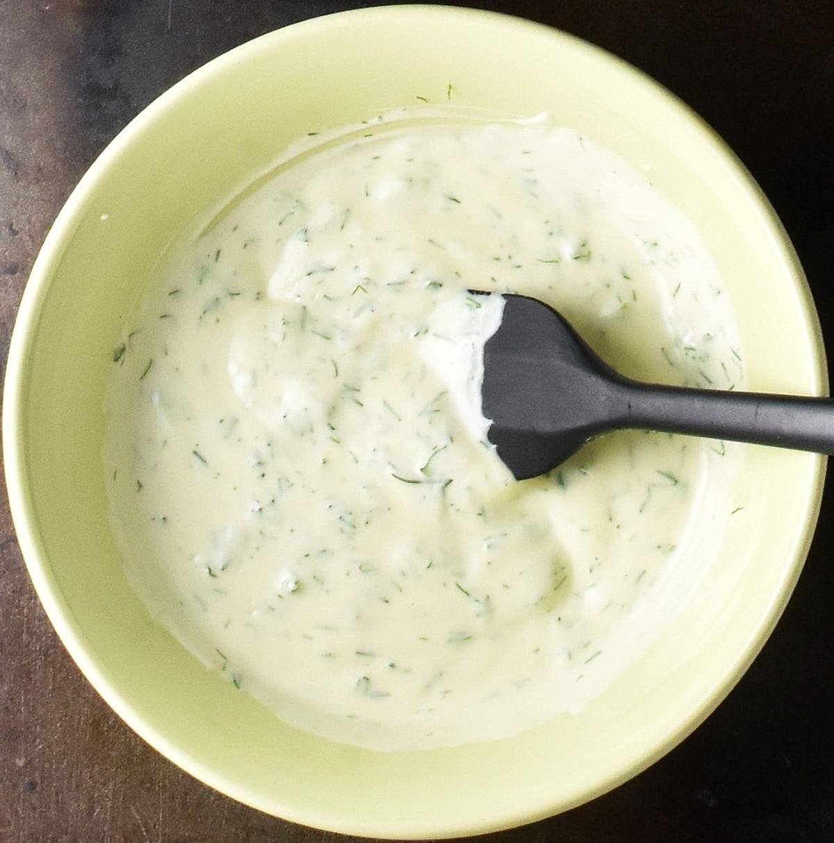 Top down view of creamy dill dressing in yellow bowl with black spoon.