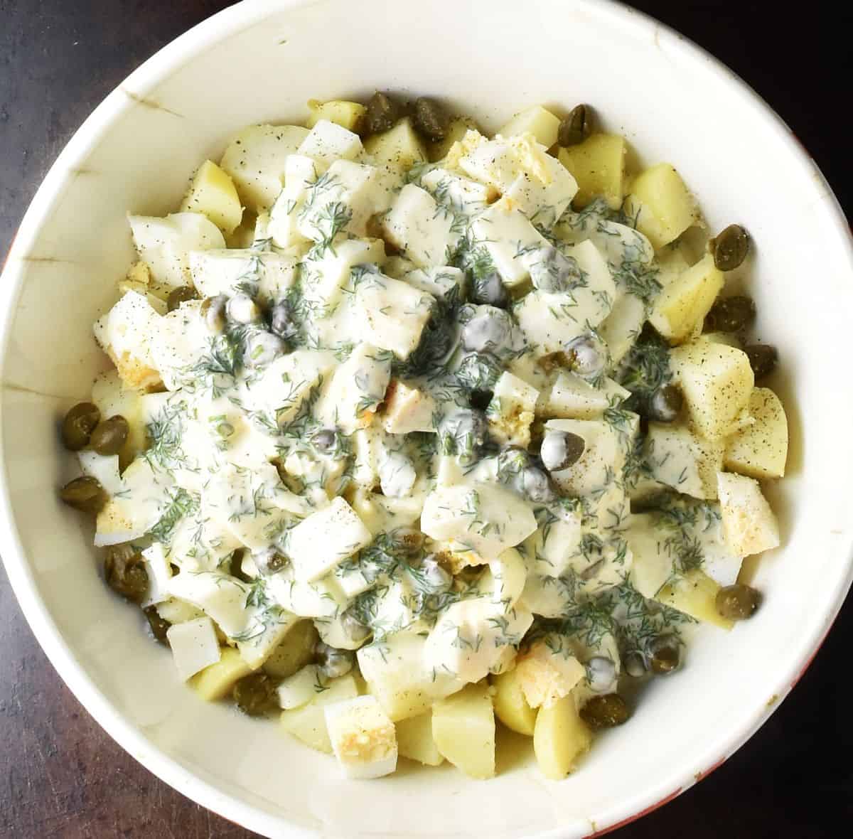 Potato dill salad ingredients with creamy dressing in yellow bowl.