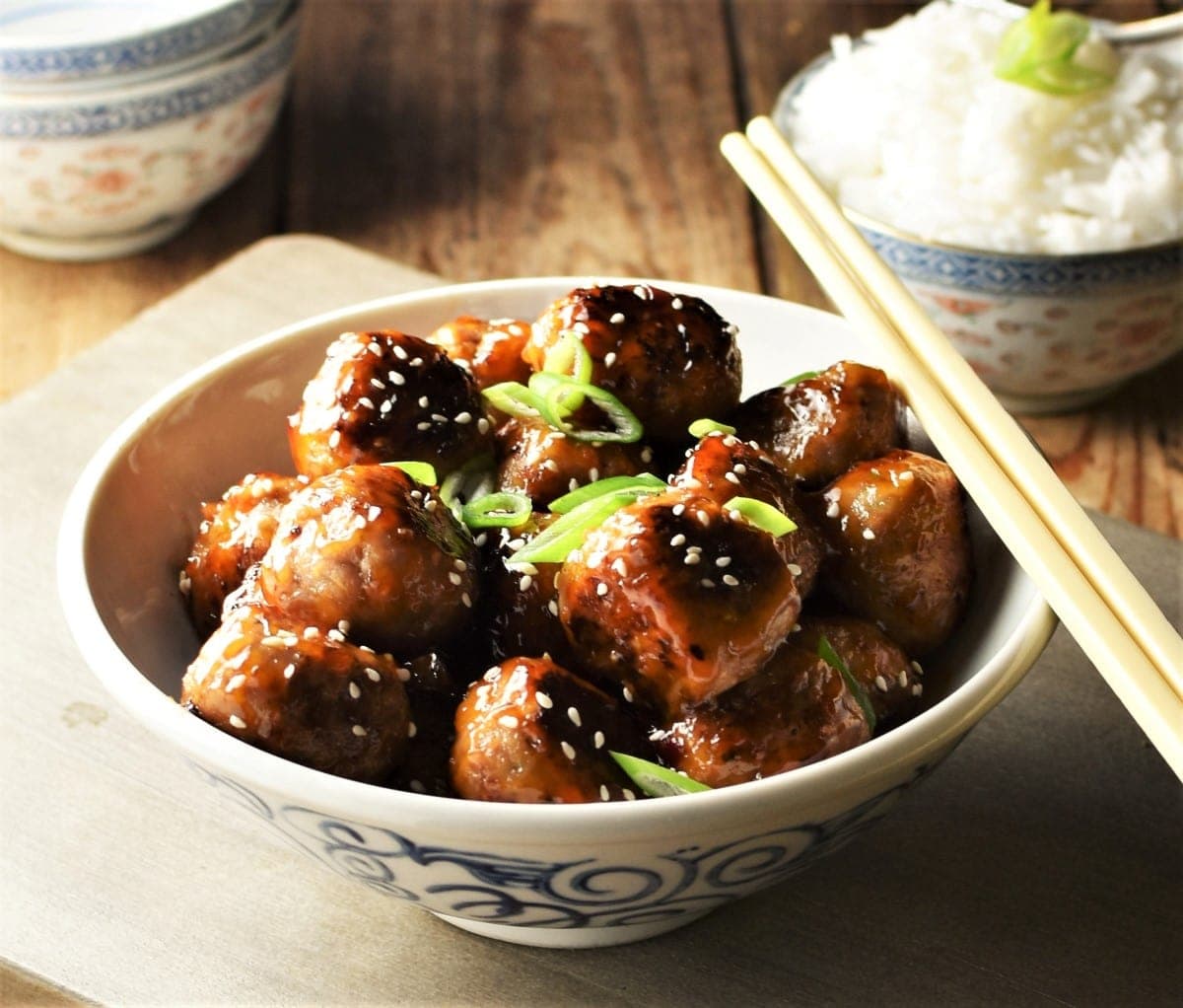 Close-up view of meatballs with glaze in bowl with chopsticks and rice in background.
