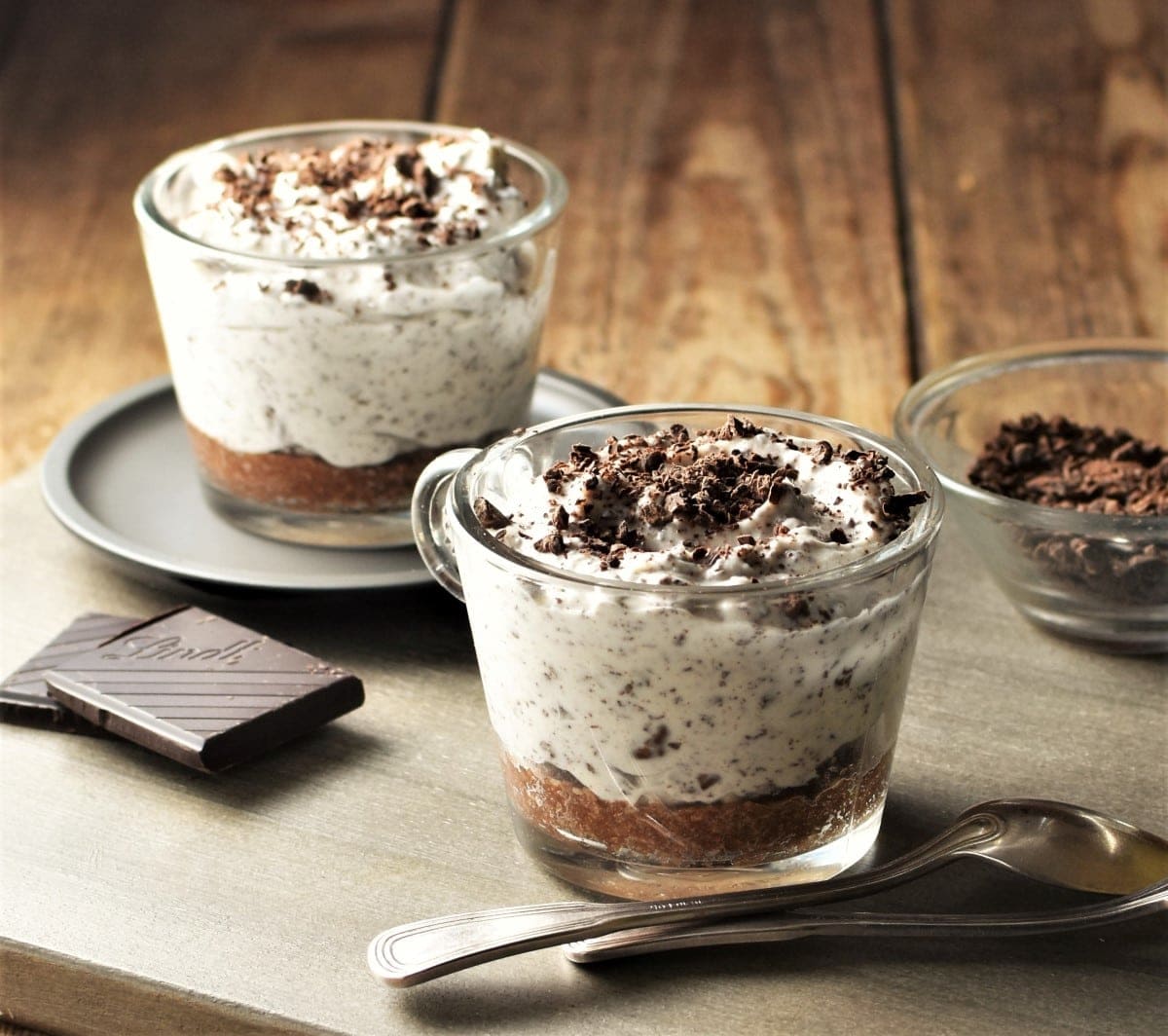 Side view of 2 cheesecake cups with chocolate shavings and spoons in background.