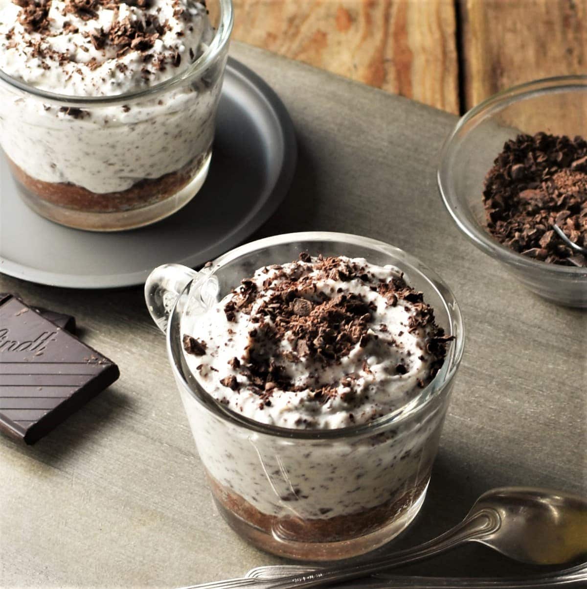 Close-up view of 2 stracciatella cheesecake cups with chocolate and spoons in background.
