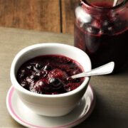 Side view of fruit compote in white bowl with spoon on top of saucer with compote in jar in background.