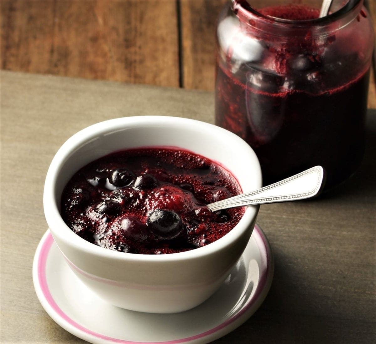 Side view of fruit compote in white bowl with spoon on top of saucer with compote in jar in background.