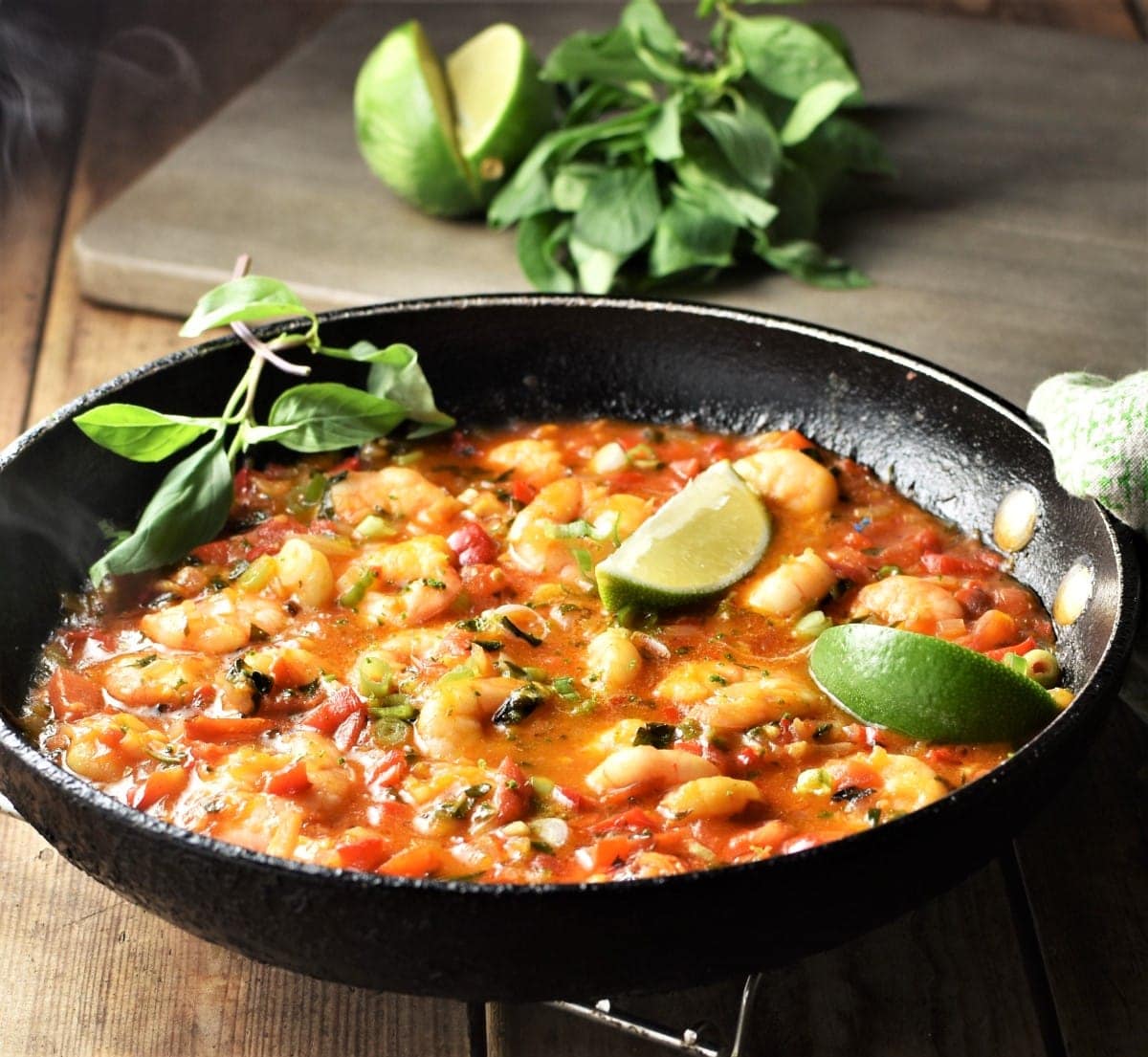 Side view of shrimp in vegetable sauce in pan with lime and herbs in background.
