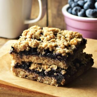 Side view of 2 blueberry bars, with white cup and blueberries in background.