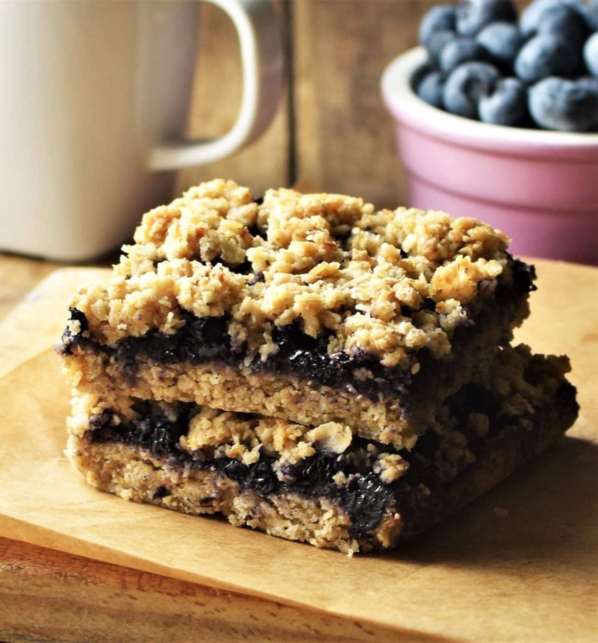 Side view of 2 blueberry bars, with white cup and blueberries in background.
