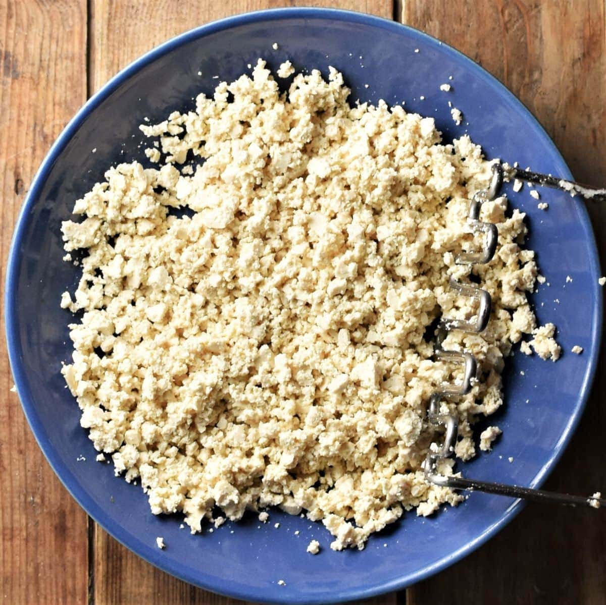 Crumbled tofu in blue bowl with potato masher.