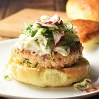 Side view of fresh salmon burgers on top of bun with slaw and buns in background.
