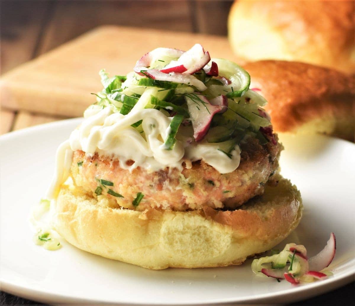 Salmon burger on top of bun with slaw and buns in background.