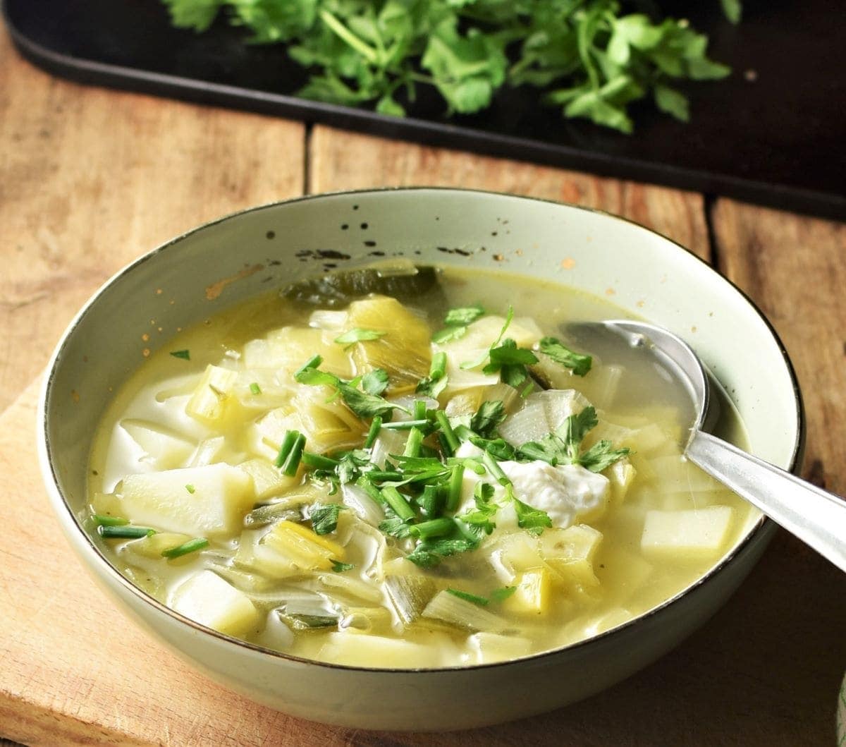 Side view of chunky leek and potato soup with herbs in green bowl with spoon.