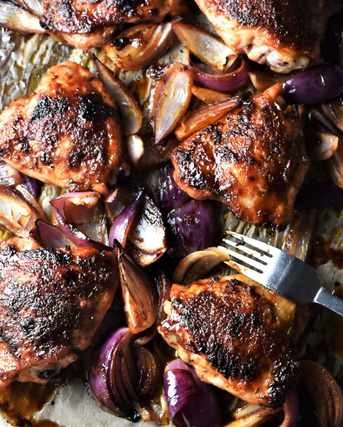 Sticky roasted chicken thighs and onions with fork on parchment.