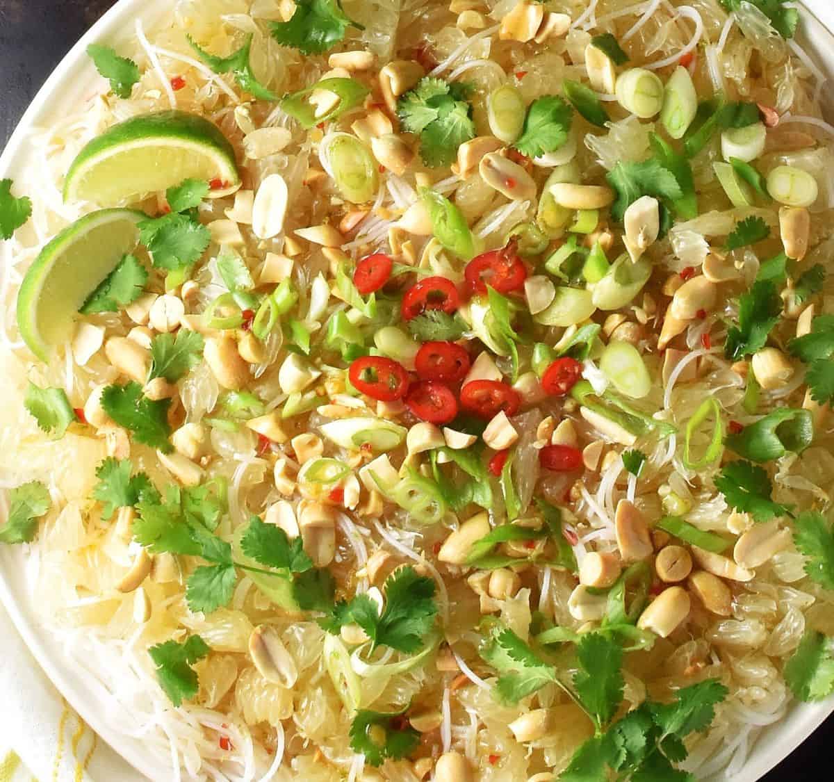 Close-up view of pomelo and noodle salad with herbs and lime wedges on white plate.
