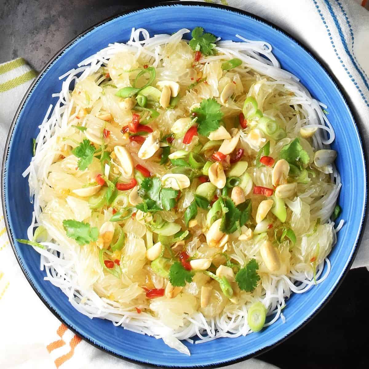 Top down view of pomelo salad with noodles, herbs and nuts in blue bowl.