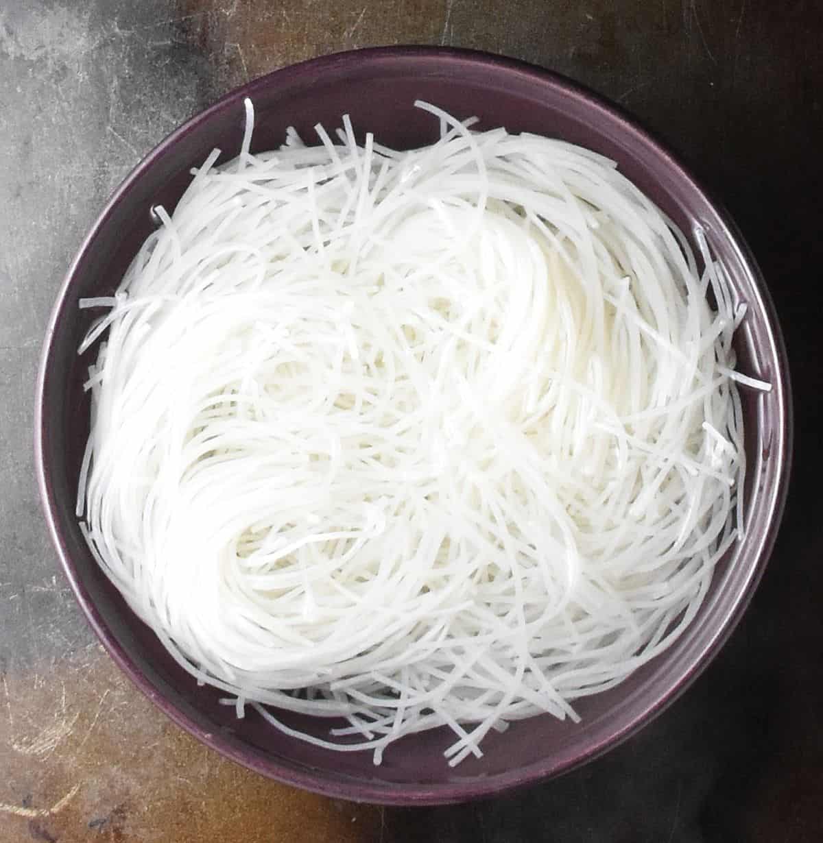 Rice noodles soaking in water in purple bowl.