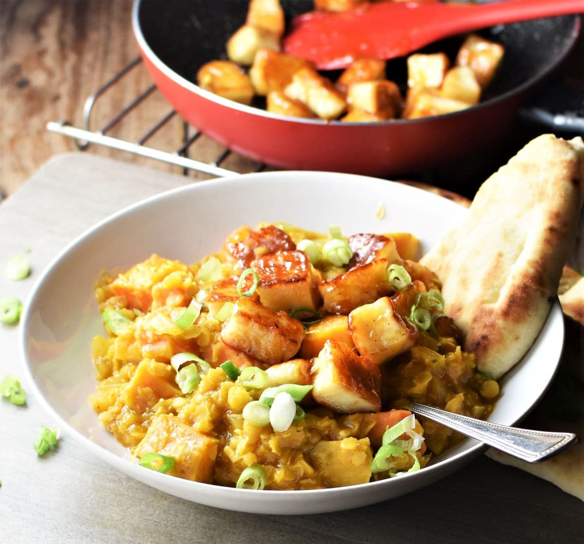 Side view of dhal with sweet potato and halloumi and naan in white bowl with spoon and halloumi cubes in pan in background.