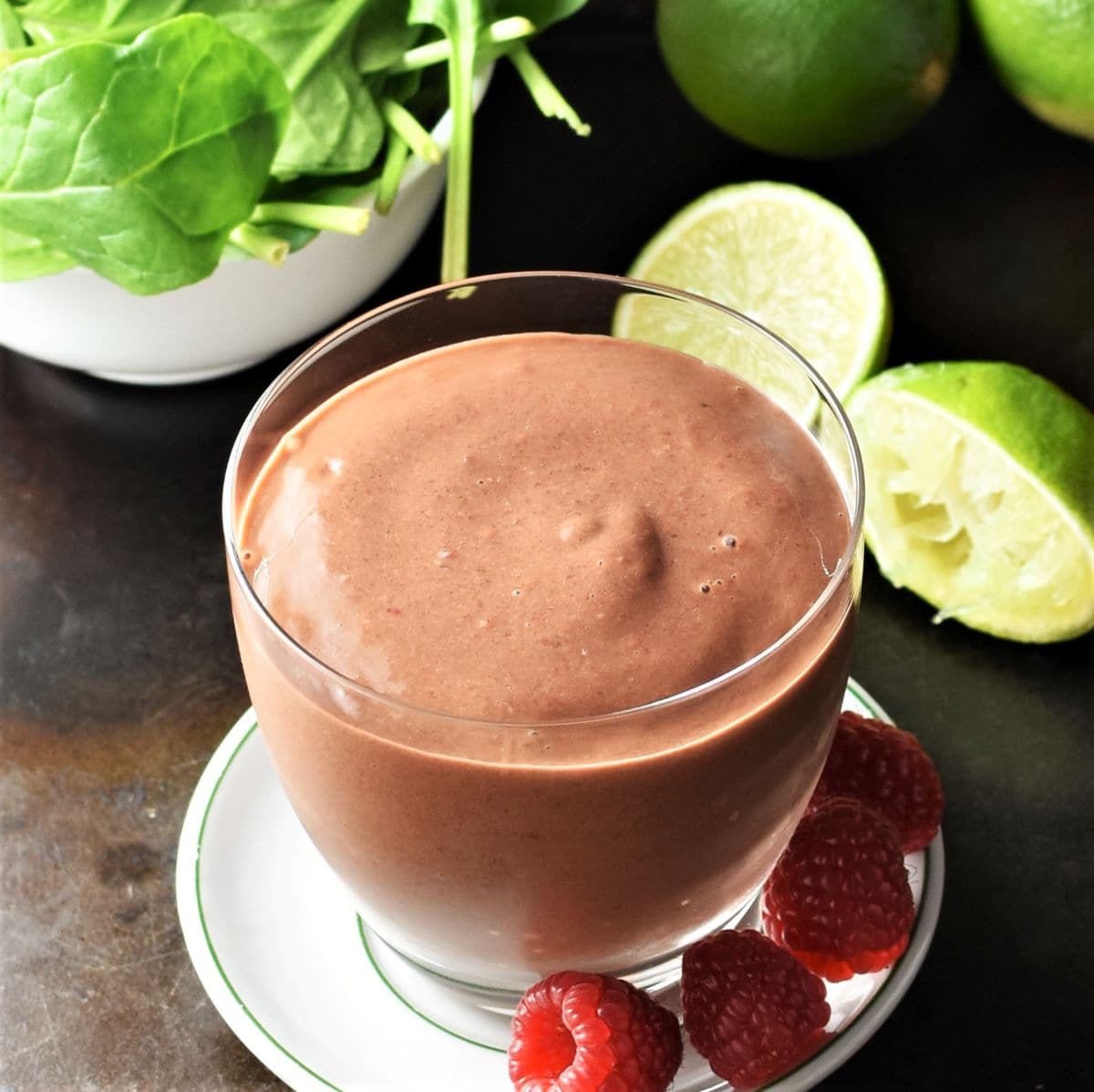 Close-up view of smoothie in glass on top of saucer with raspberries and limes in background.