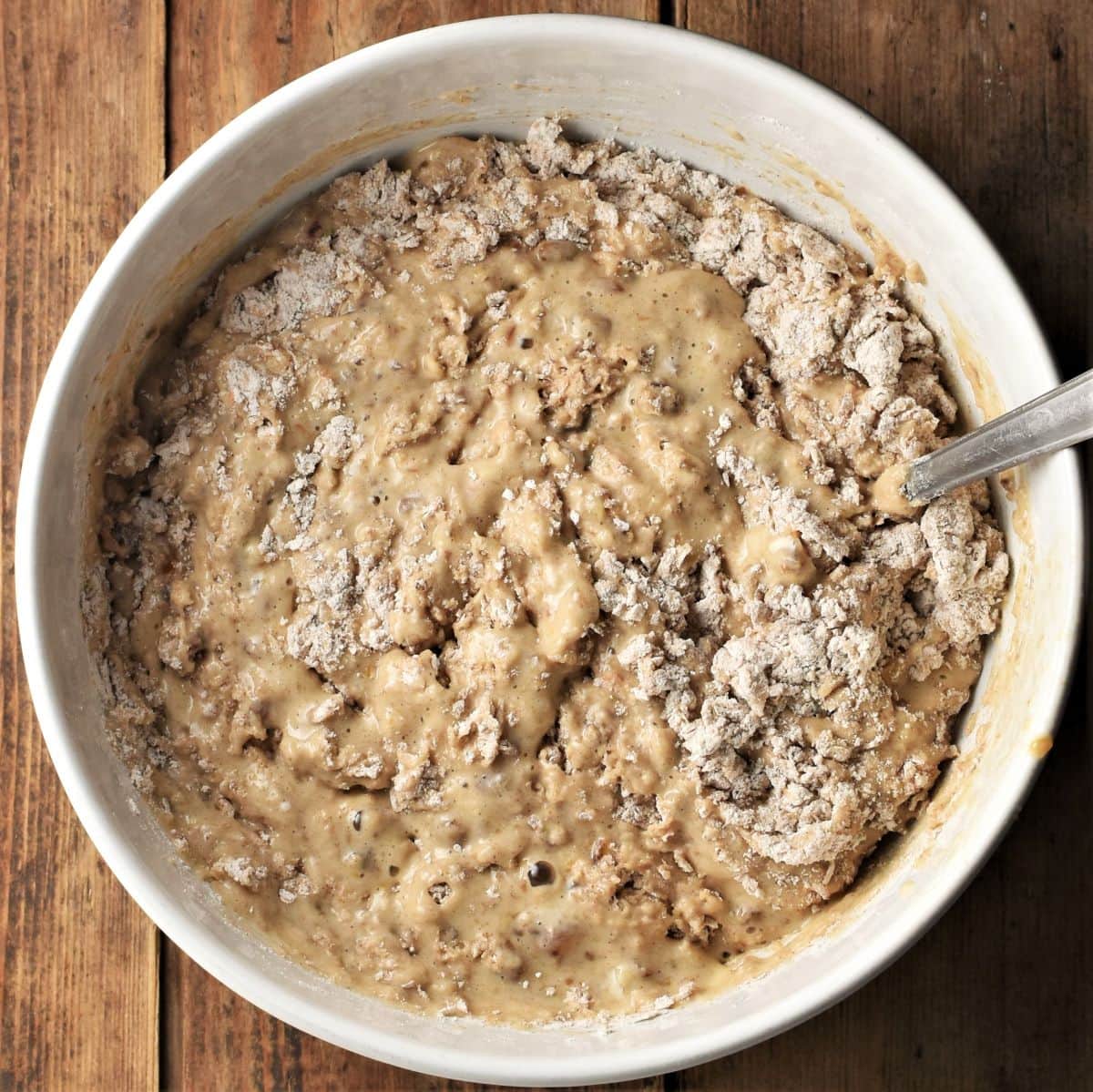 Combining banana and date bread wet and dry ingredients in white bowl with spoon.