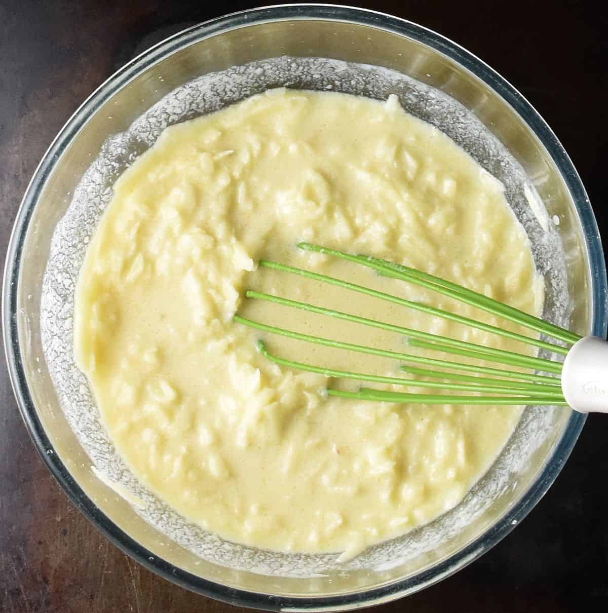 Wet mixture with grated apple in large, glass bowl with green whisk.