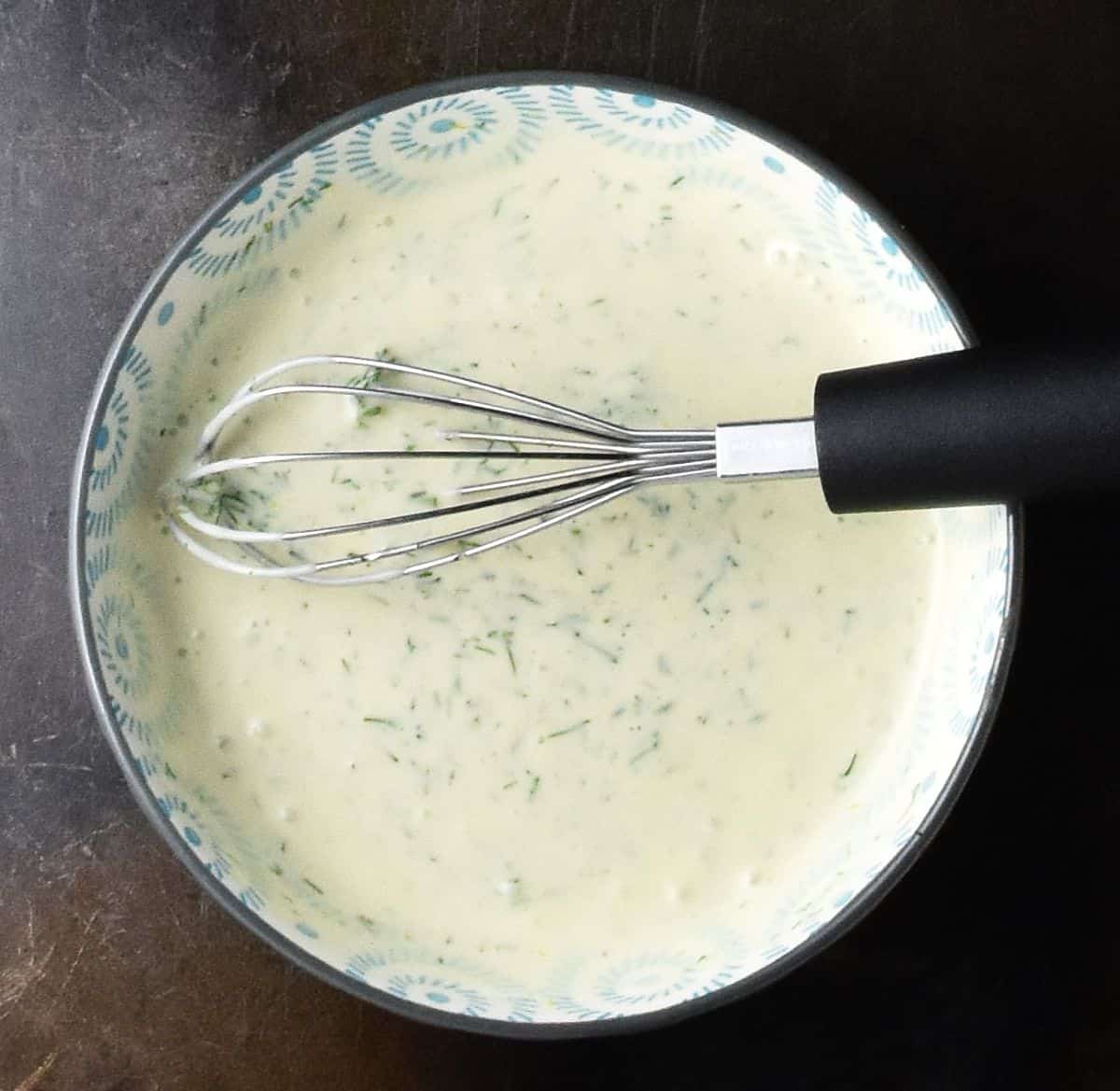 Top down view of creamy dressing with dill in blue bowl with whisk.