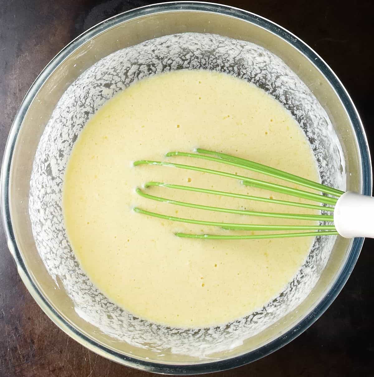Creamy pancake wet mixture with green whisk in glass bowl.