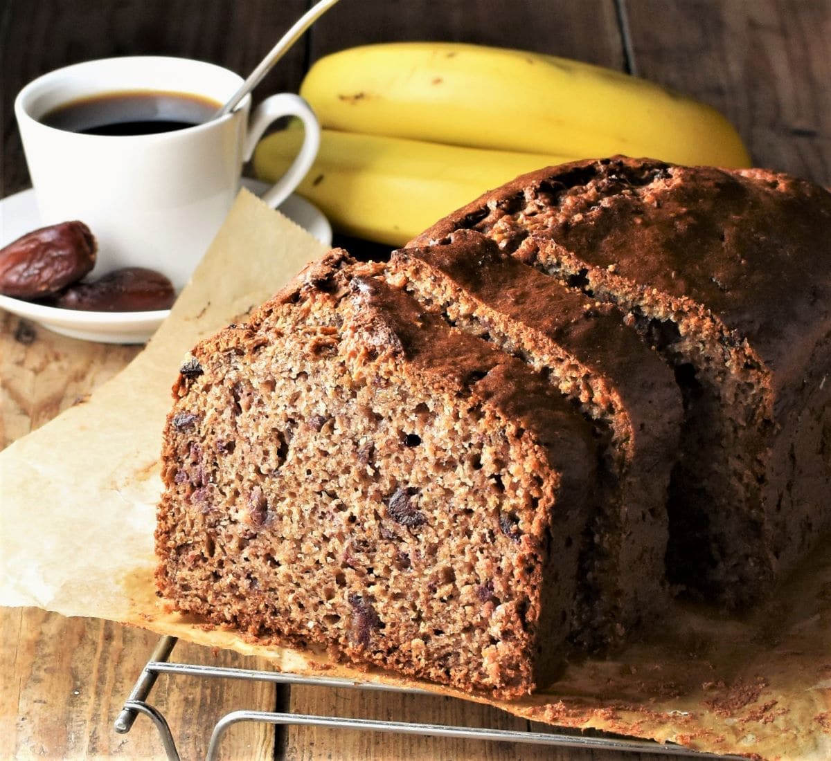 Side view of sliced banana bread on top of parchment with coffee cup and bananas in background.