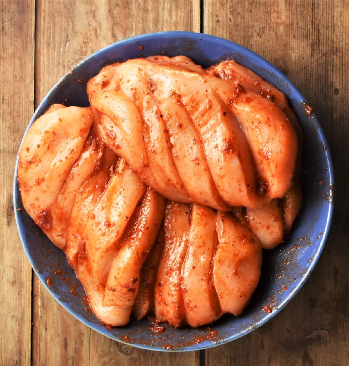 Raw hasselback chicken pieces coated in harissa paste in blue bowl.