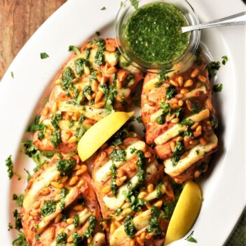 Baked hasselback chicken breasts with halloumi, herb sauce and lemon wedges in white dish.