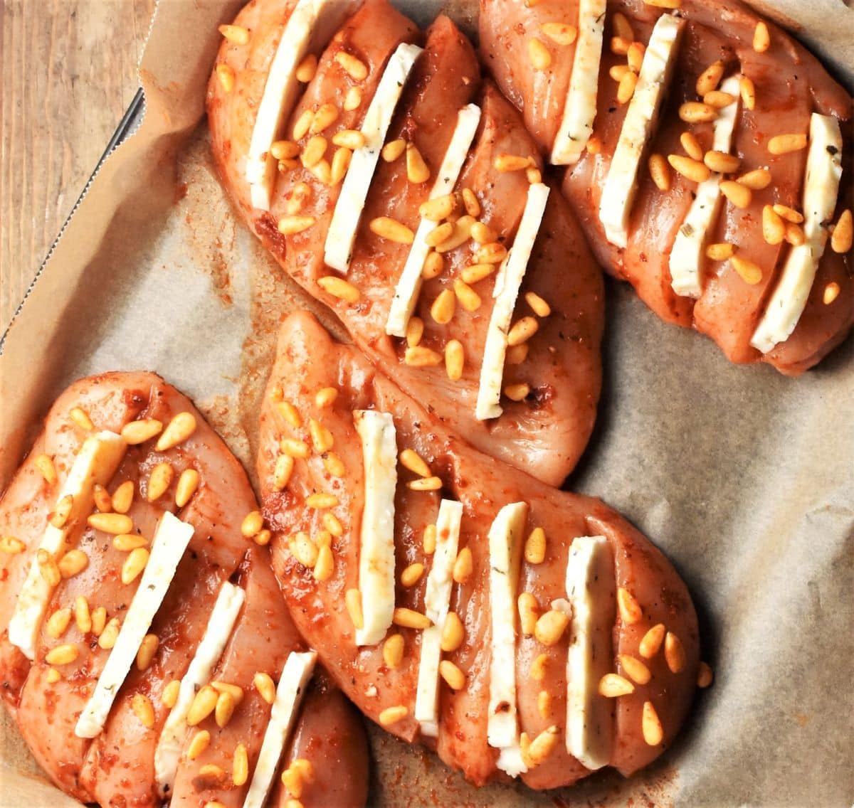 4 raw hasselback chicken breasts with halloumi and pine nuts on top of baking sheet.