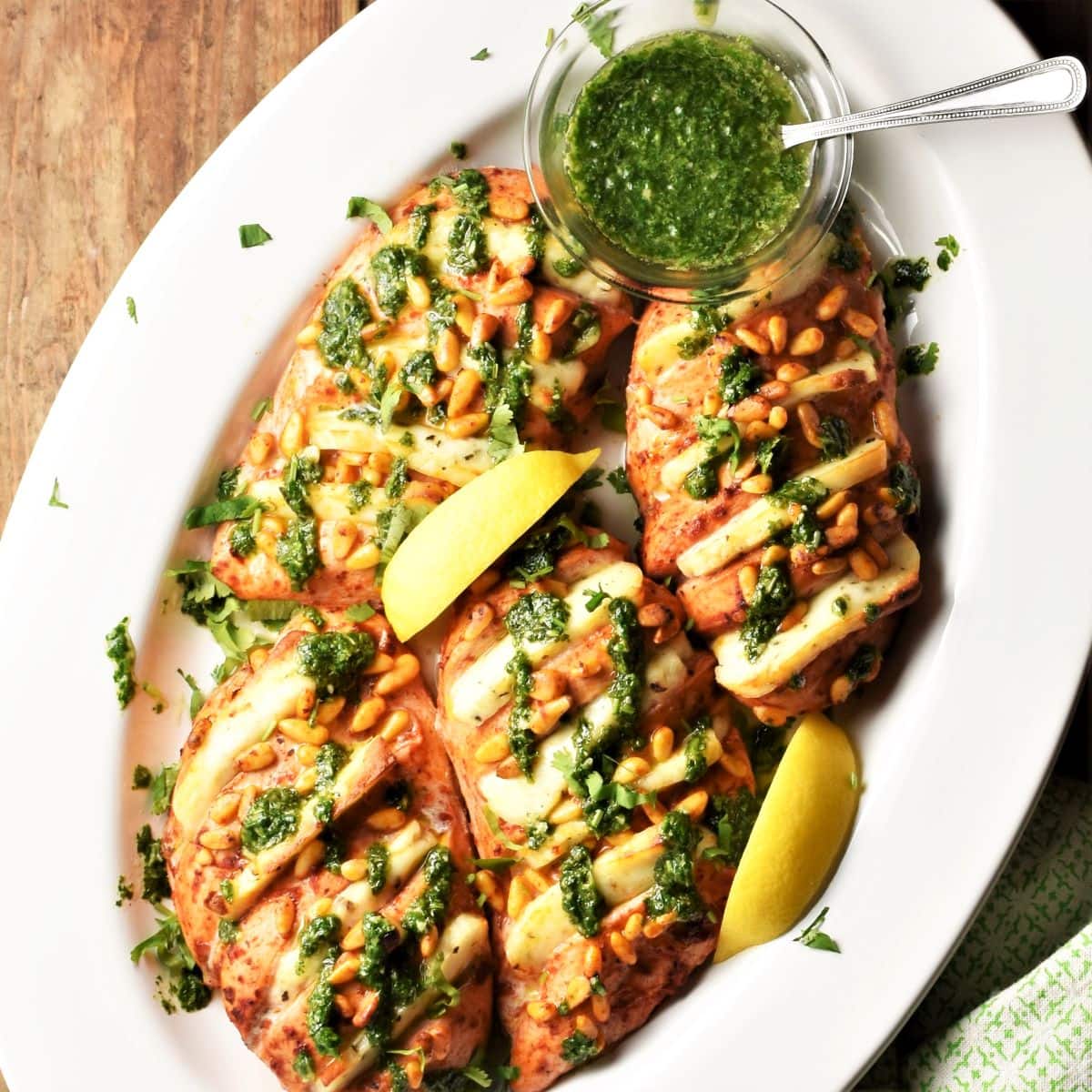 Baked hasselback chicken with halloumi, lemon wedges and herb sauce in oval white dish.