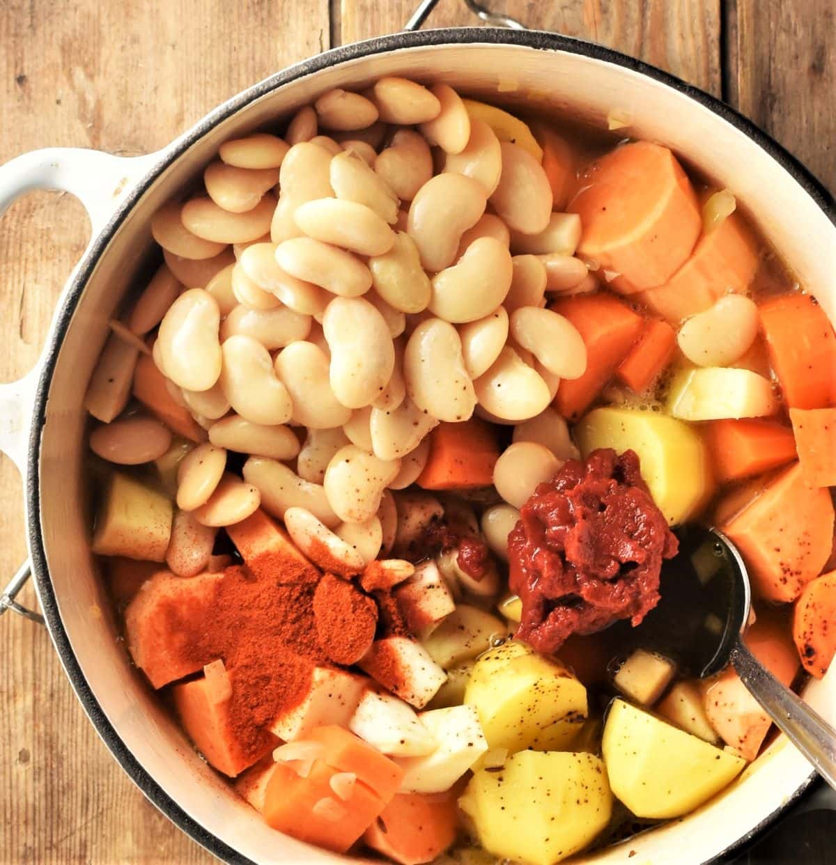 Assembling root vegetables, beans and spices in large pot with spoon.