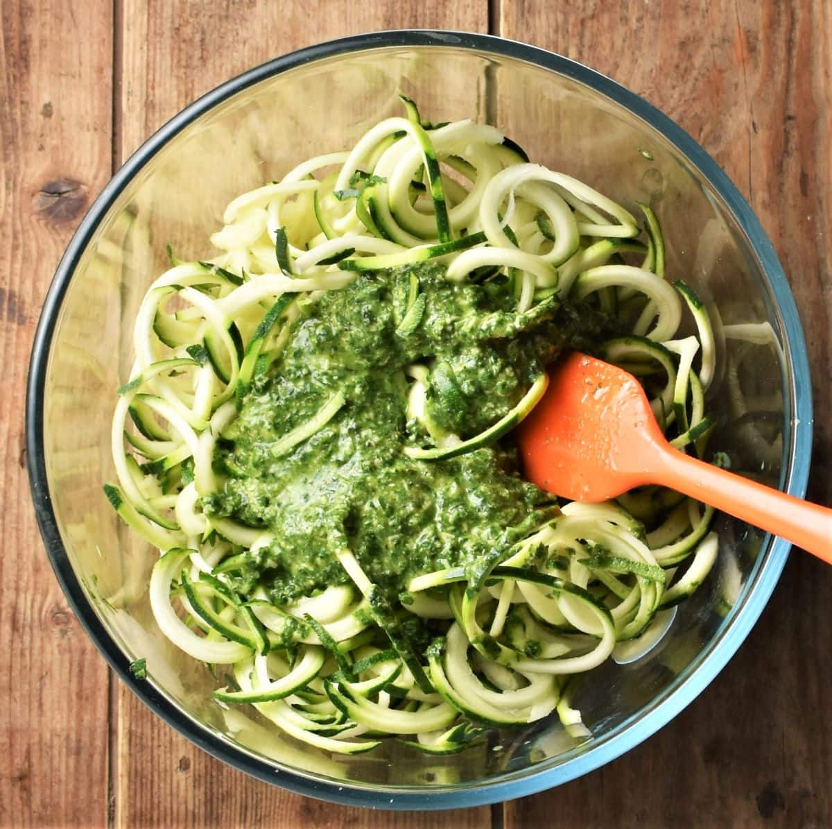 Zucchini noodles with pesto sauce in mixing bowl with red spatula.