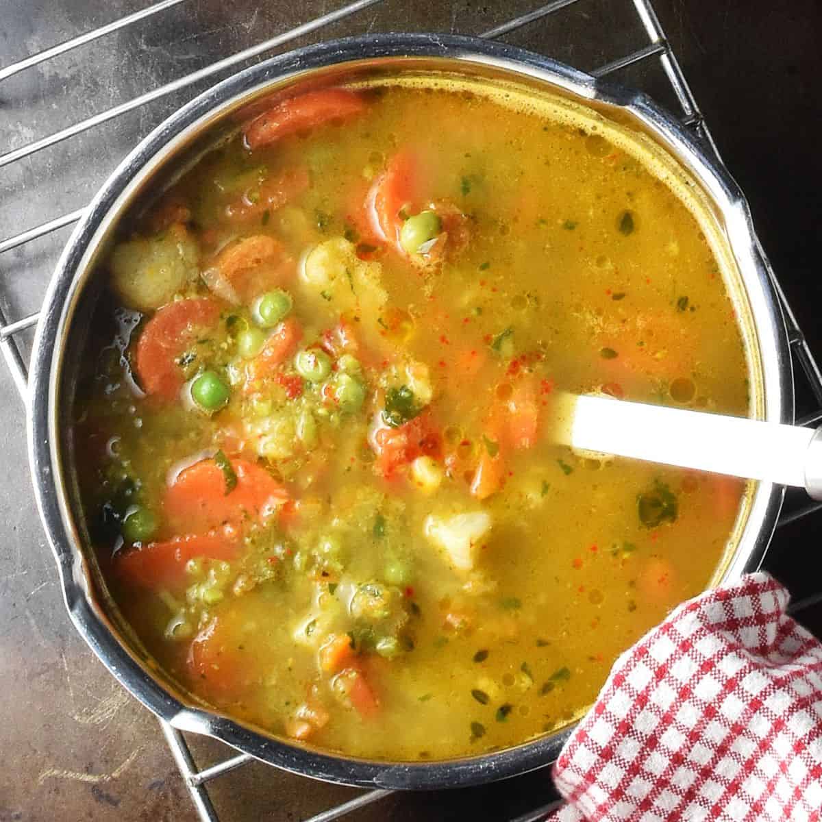 Top down view of frozen vegetable soup with chunks of veg in metal pot with ladle.