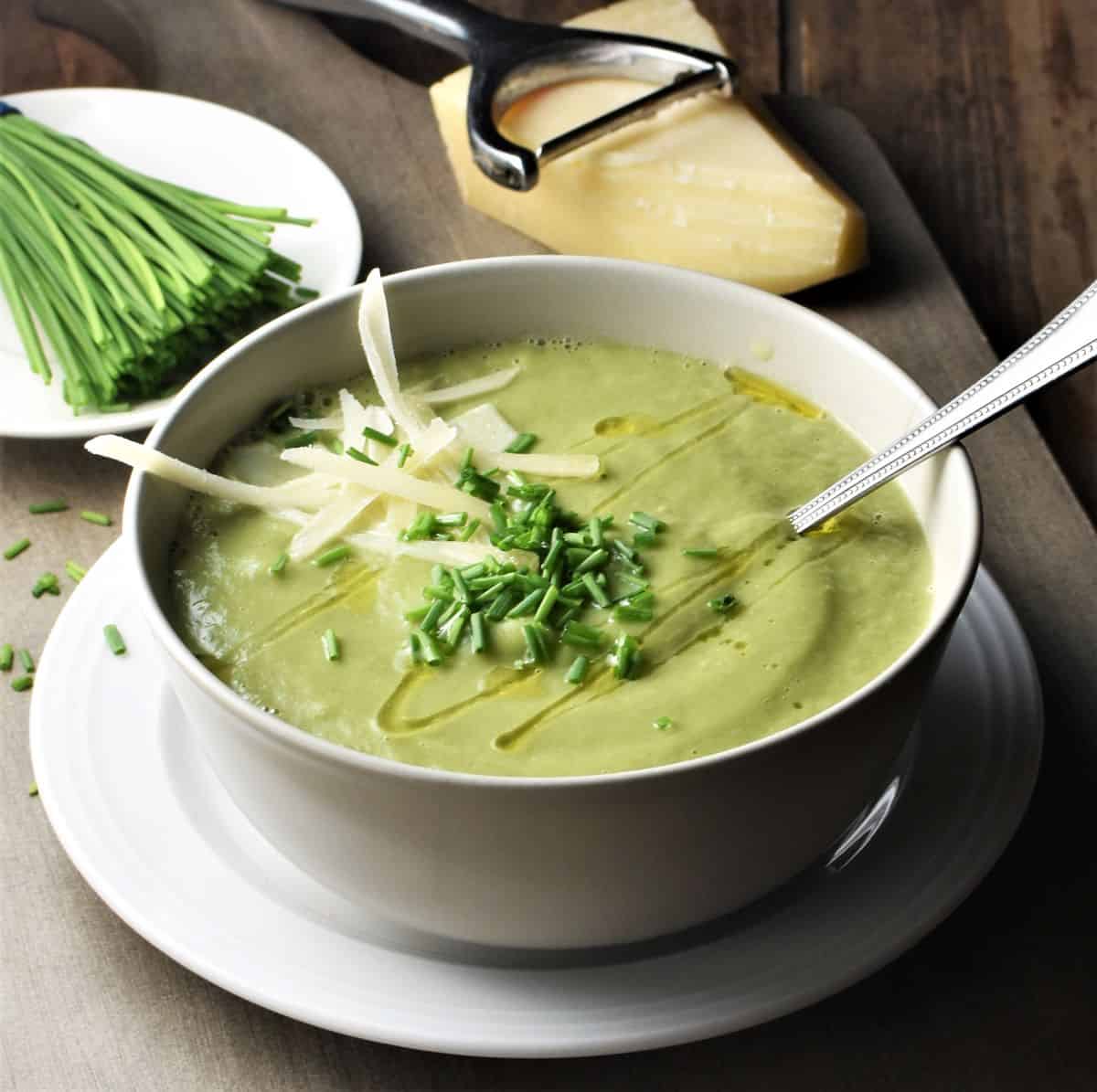 Side view of creamy fennel soup in white bowl with spoon, chives, vegetable peeler and cheese in background.