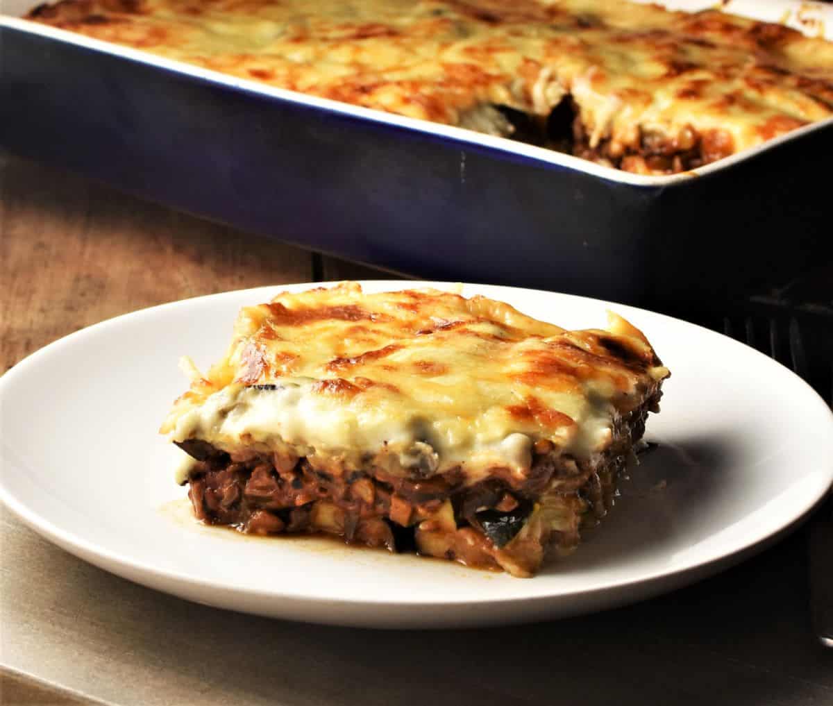 Side view of lentil mushroom moussaka slice on top of white plate with moussaka in background.
