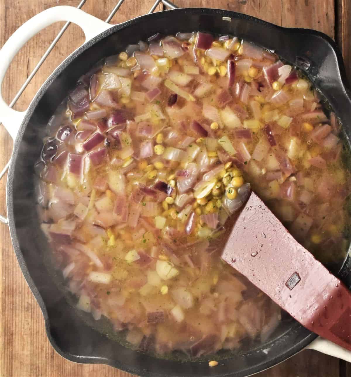 Cooking onion and green lentils with stock in skillet with red spatula.