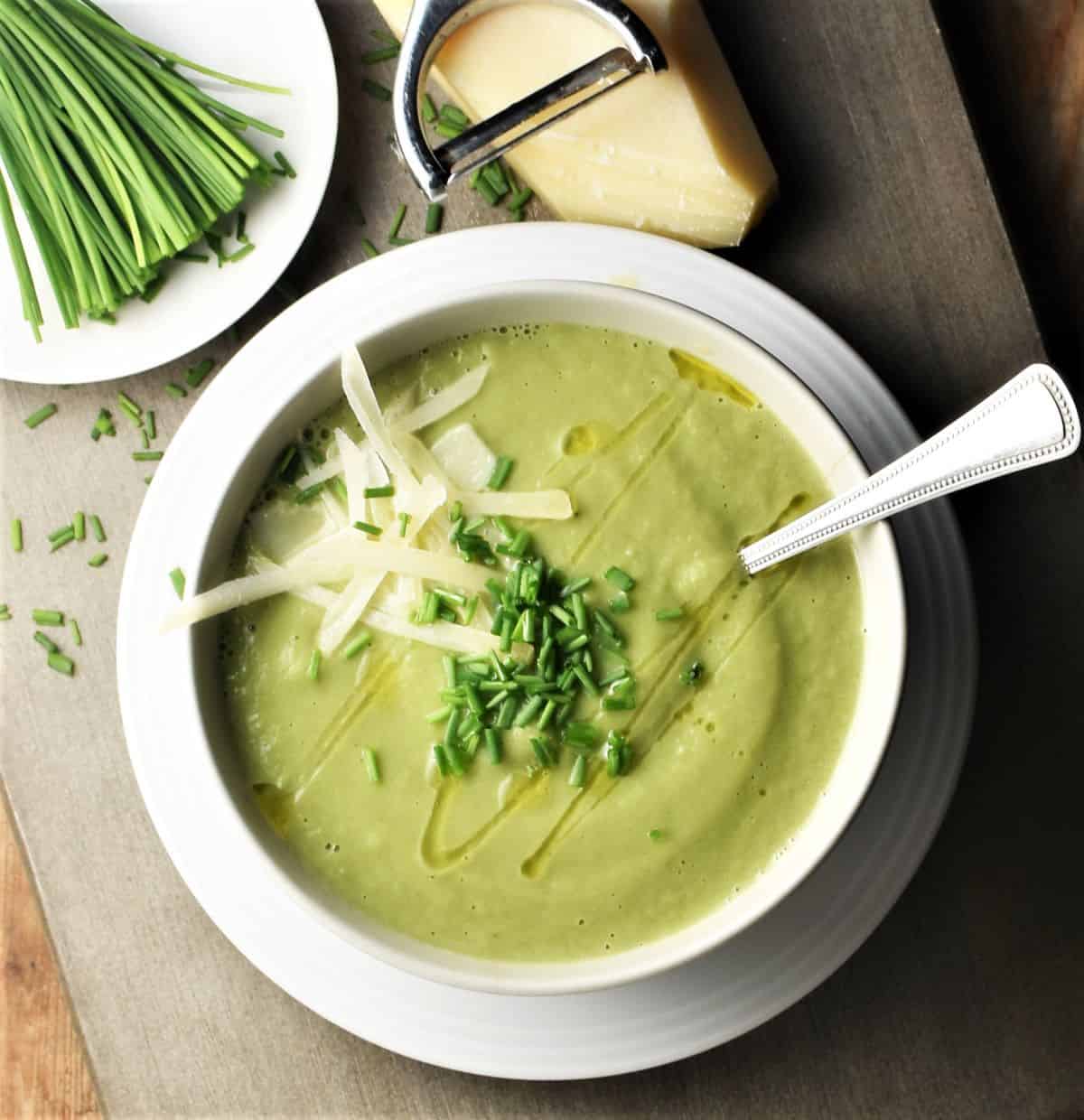 Top down view of creamy fennel soup in white bowl with spoon, chives and cheese in background.