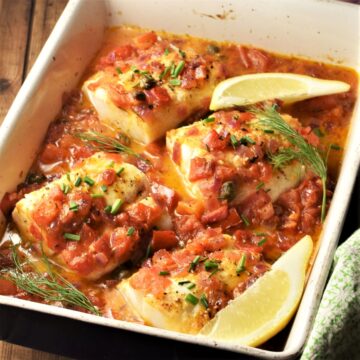 Side view of 4 cod pieces in tomato sauce in casserole dish with lemon wedges.