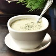 Side view of creamy dill mustard sauce in white bowl with spoon on saucer.