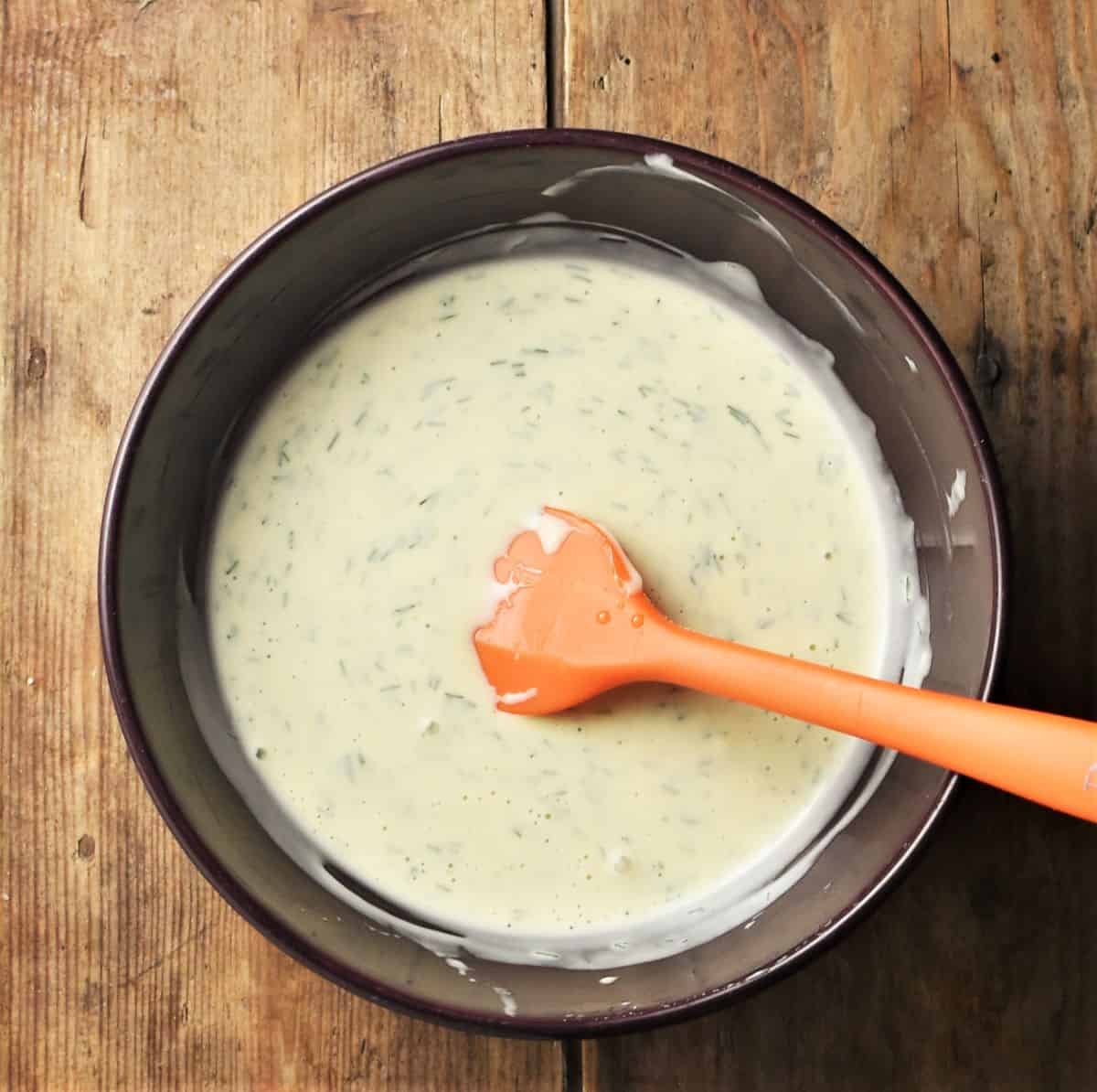 Top down view of creamy dill sauce in purple bowl with orange spatula.