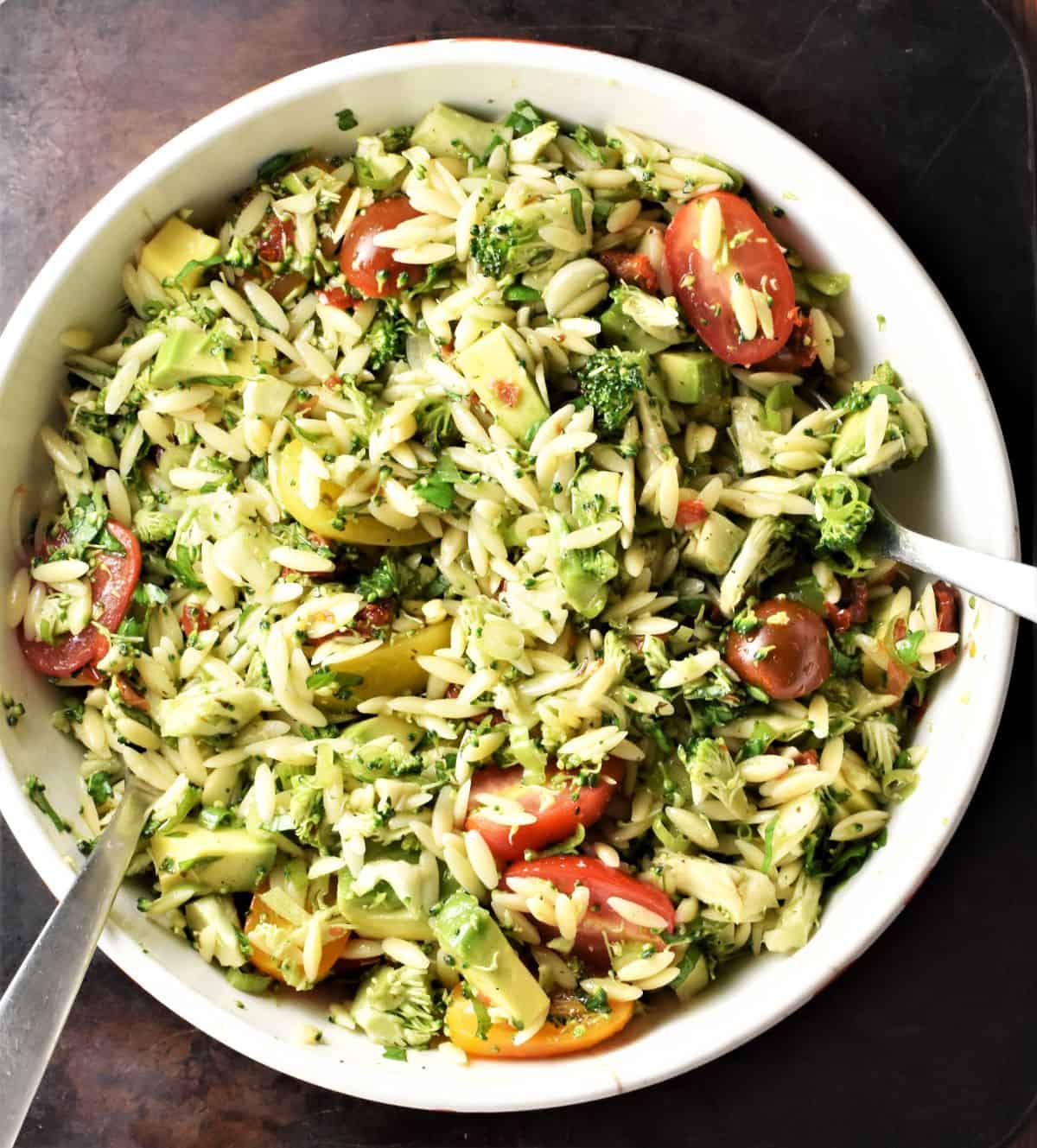 Broccoli salad with pasta and tomatoes in large white bowl with spoons.
