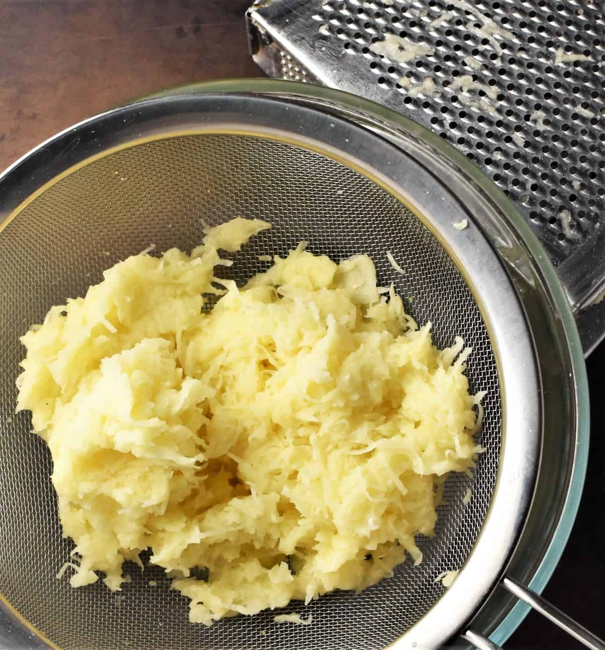 Shredded potato in sieve with grater in background.