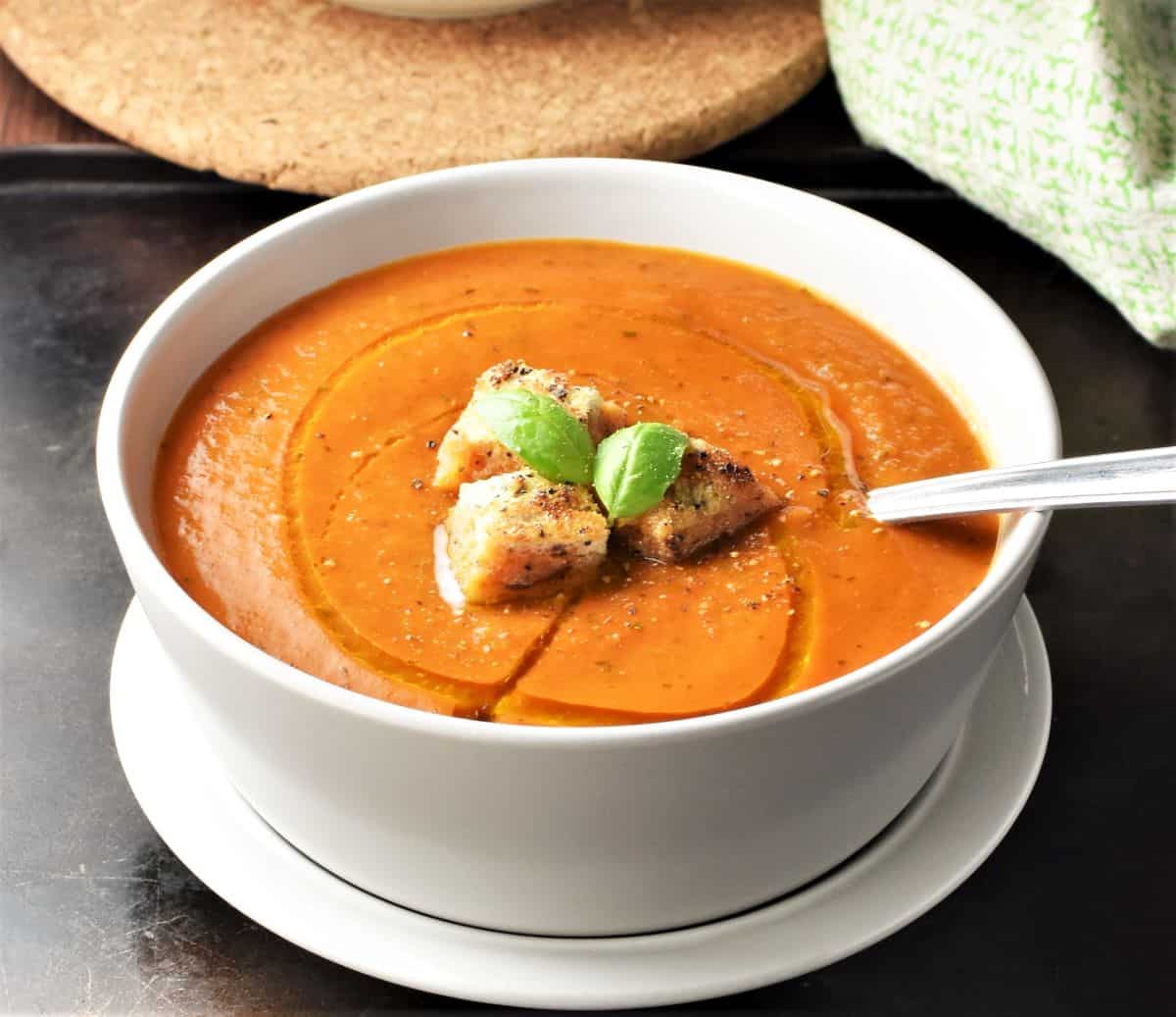 Side view of creamy tomato soup with croutons and spoon in white bowl.
