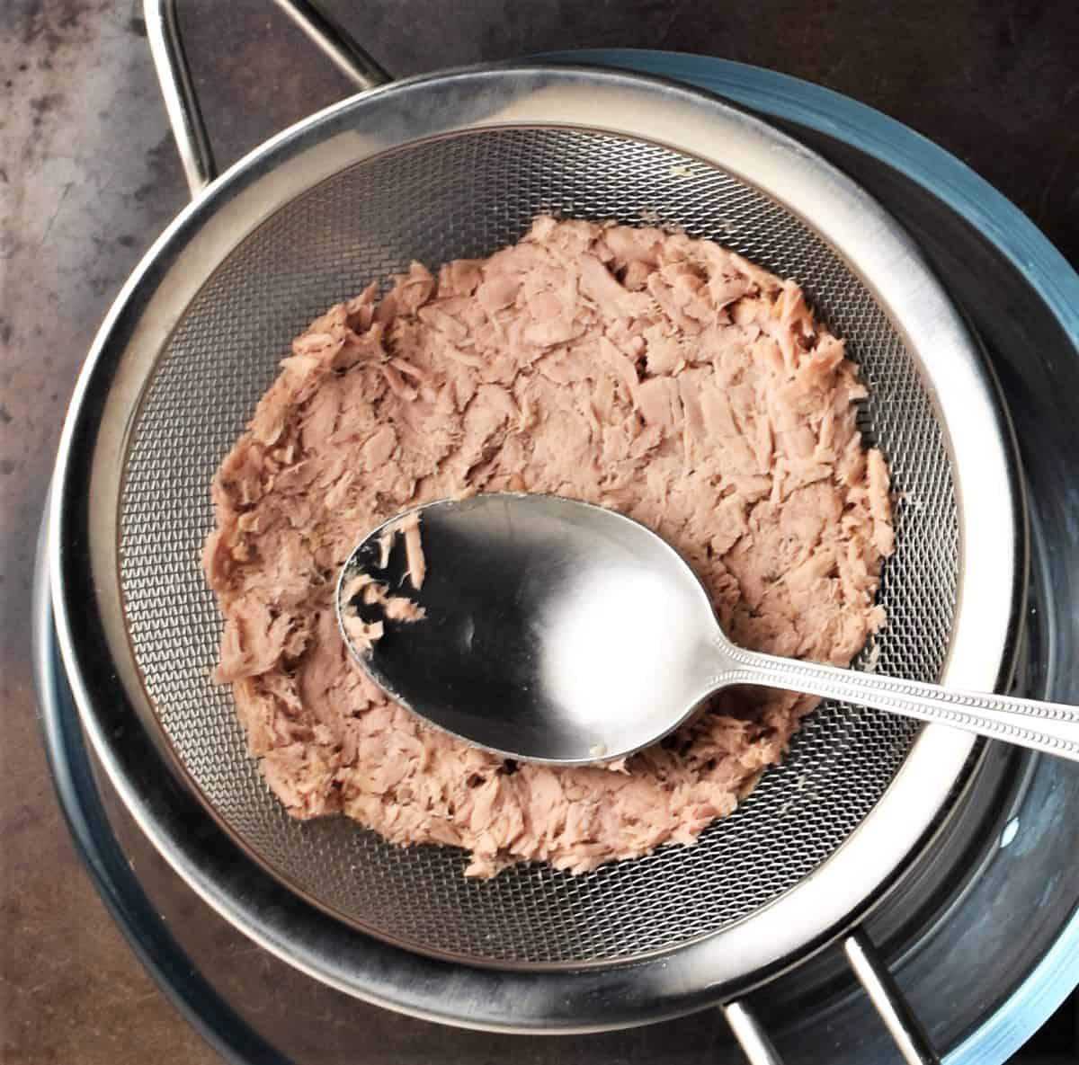 Squash tuna in sieve with spoon over bowl.