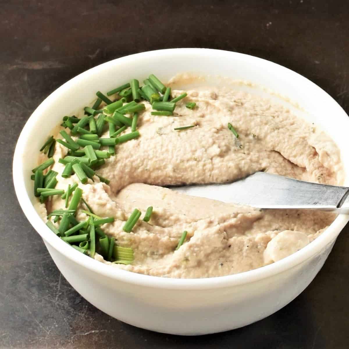 Side view of creamy tuna spread with chives and knife in white bowl.