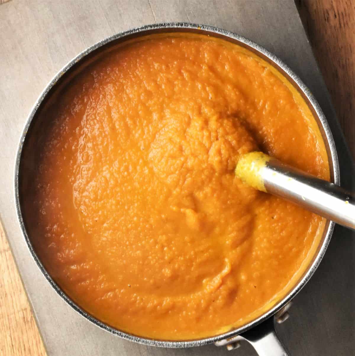 Pureeing pumpkin carrot soup with stick blender in pot.