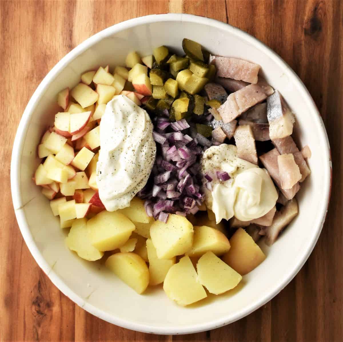 Assembled ingredients in white bowl for Polish herring potato salad.